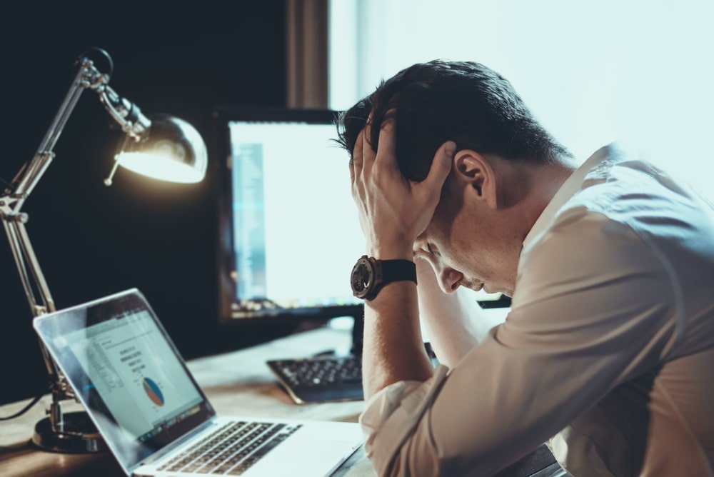 Um homem, aparentemente estressado ou sobrecarregado, está sentado em uma mesa com as mãos na cabeça, em frente a um laptop e um monitor. A iluminação é baixa e uma luminária de mesa acesa adiciona foco à cena, representando os desafios na criação de Reels para empresas.