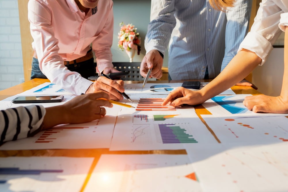 Equipe de negócios reunida em torno de uma mesa repleta de documentos e gráficos impressos, apontando para dados para melhorar o engajamento do cliente.