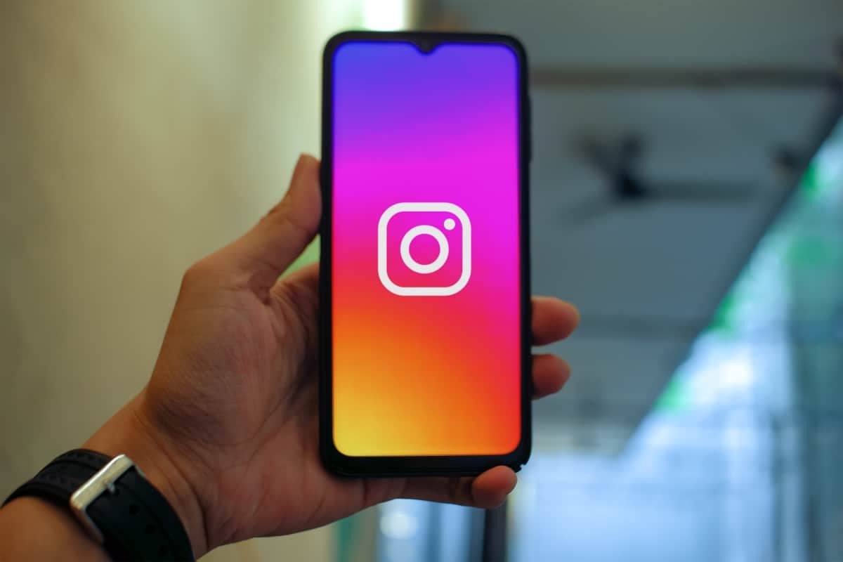 Uma mão segura um smartphone que exibe a tela de abertura do aplicativo Instagram, com o logotipo da câmera centralizado em um fundo de gradiente roxo, rosa, laranja e amarelo. A imagem simboliza o início ou o fim do uso do aplicativo e a decisão de gerenciar o tempo de tela com recursos como o modo silencioso.