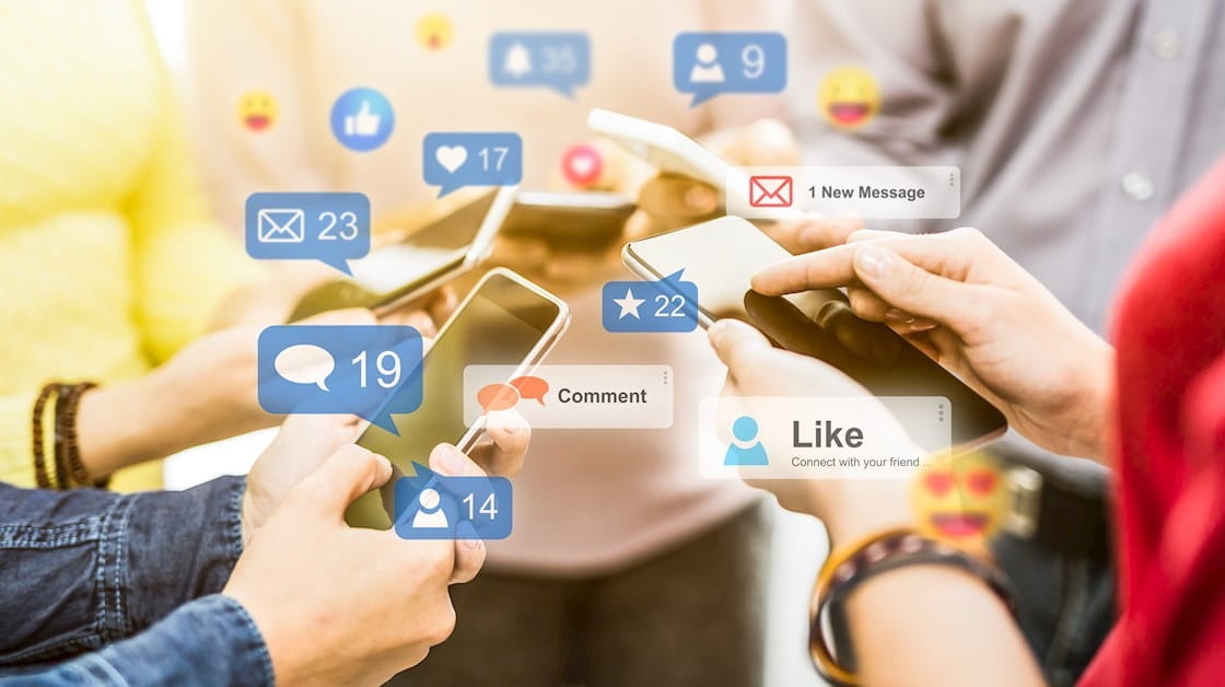 Uma imagem com foco em smartphones. As mãos de várias pessoas seguram os dispositivos, e sobre as telas flutuam ícones de redes sociais, como balões de fala, curtidas e emojis. A cena sugere a intensa interação social e o engajamento digital, com a mensagem principal de vender no Instagram sendo comunicada através dos elementos visuais que representam a comunicação e o alcance nas mídias sociais.