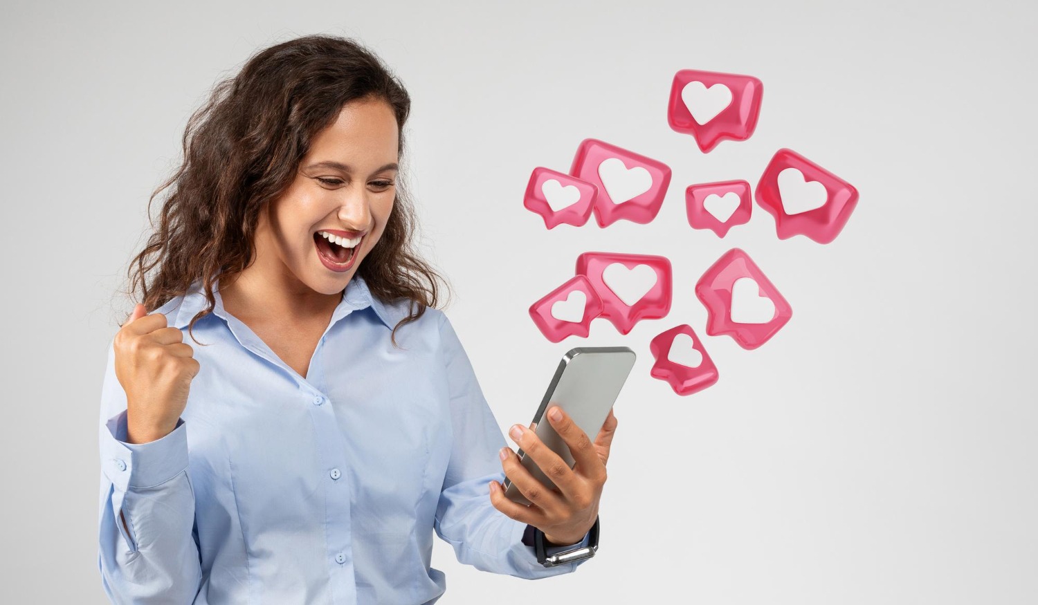 Uma mulher jovem e de cabelo escuro, vestindo uma camisa social azul clara, segura um smartphone com as duas mãos e sorri. Ela olha para a tela com uma expressão de surpresa e alegria, com uma das mãos fechada em punho, em um gesto de comemoração. Ícones 3D de corações, representando curtidas e engajamento, flutuam ao lado do smartphone, em diversas formas e tamanhos. A imagem sugere a sensação de sucesso e satisfação que se tem ao vender no Instagram e ver o engajamento crescer.