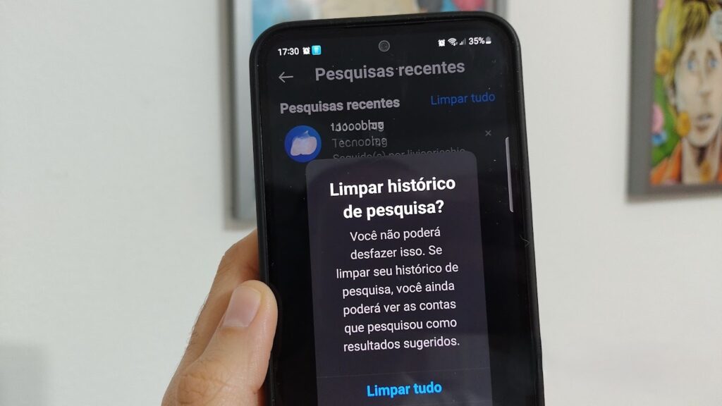 Uma foto em close-up de um smartphone sendo segurado por uma mão, exibindo o aplicativo do Instagram na tela de "Pesquisas recentes". Um pop-up escuro de confirmação aparece, perguntando "Limpar histórico de pesquisa?" e alertando que a ação não pode ser desfeita. No pop-up, a opção "Limpar tudo" está visível na parte inferior, indicando o passo final para o usuário apagar sugestões e o histórico de busca.