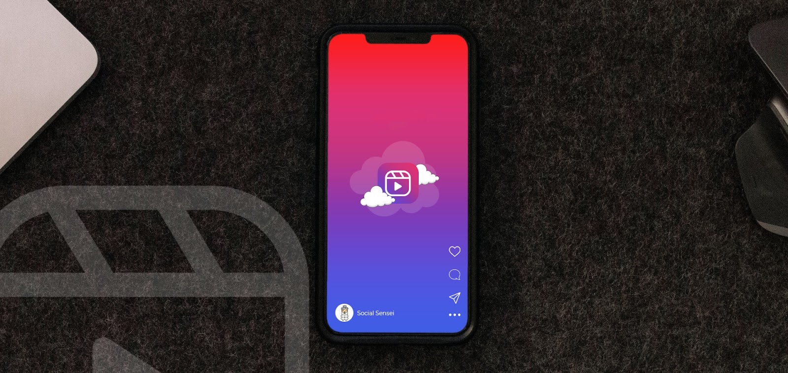 Um smartphone está posicionado sobre uma superfície escura e texturizada. Na tela, um fundo com gradiente de vermelho e roxo exibe um ícone de nuvem com o logo do Reels no centro. Partes de um laptop e um mouse aparecem nas laterais, representando o processo de criação de Reels para empresas.