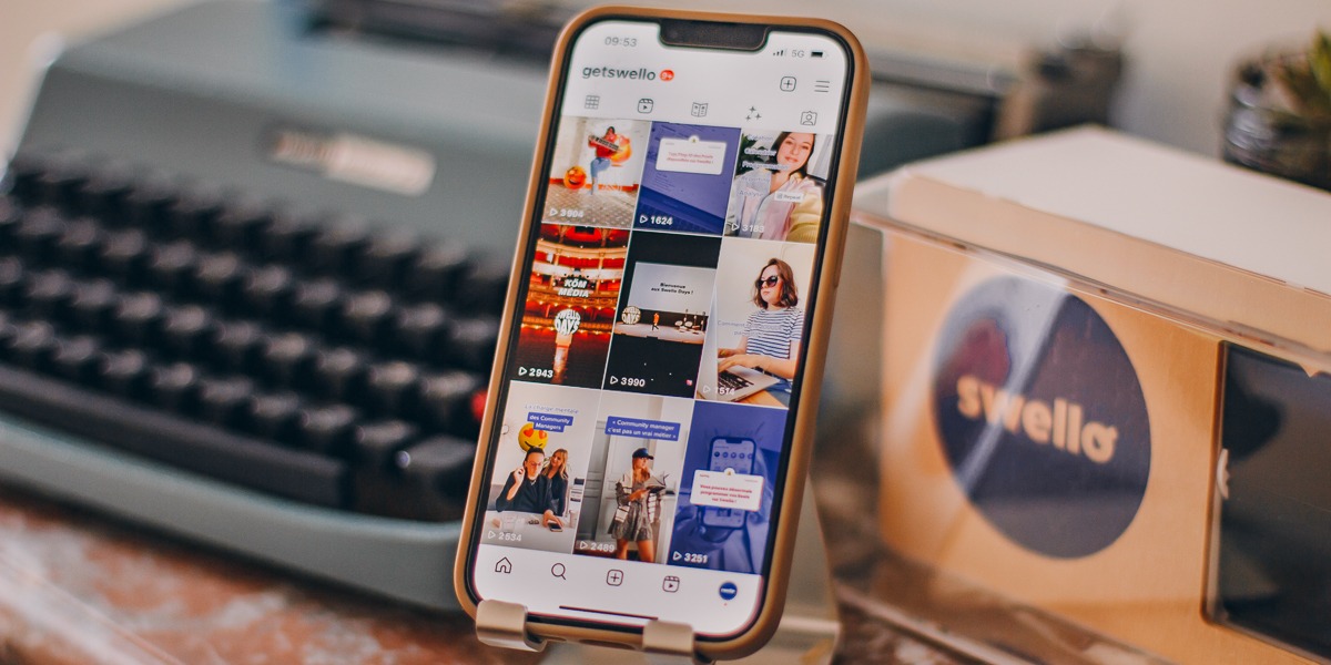 Um smartphone está em um suporte, com a tela exibindo uma galeria de vídeos do Instagram Reels, com diversas miniaturas e contagem de visualizações. Ao fundo, uma máquina de escrever vintage e uma caixa com o logo "swello" simbolizam a gestão e estratégia de Reels para empresas.
