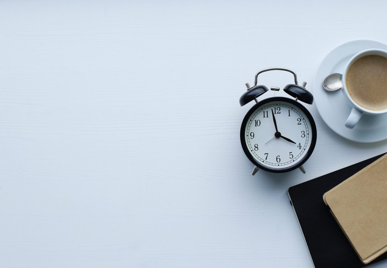 Vista superior de uma mesa branca minimalista com um despertador analógico preto, uma xícara de café com leite em pires branco e dois cadernos (um preto e um kraft). O cenário sugere rotina e produtividade, elementos importantes para planejar e otimizar o alcance dos stories.