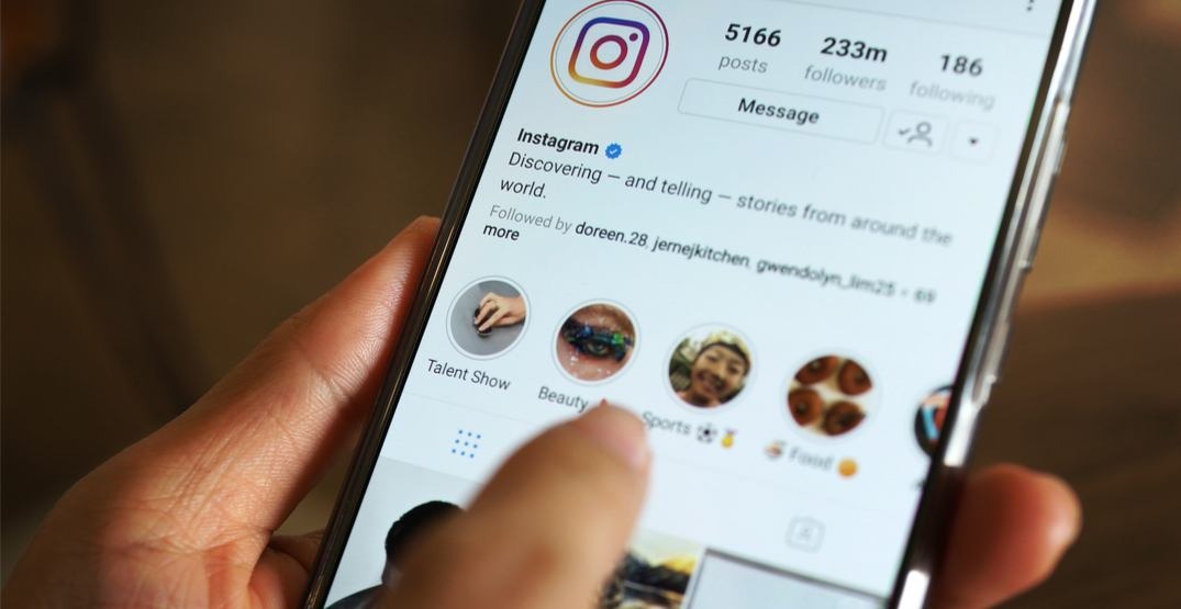 Close-up de um smartphone mostrando a tela de um perfil do Instagram com o número de seguidores em destaque, a descrição da conta (Bio no Instagram) e os círculos de Destaques (Highlights). Uma mão toca o ecrã.